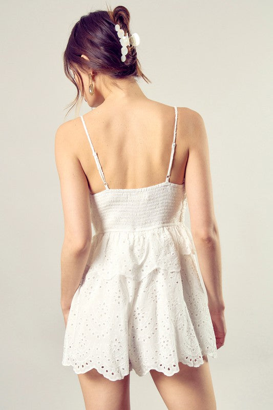 A woman wearing the Scallop Edge Front Tie-Up Romper in white, featuring a sleeveless design and eyelet texture, stands against a plain background, radiating boho style charm.