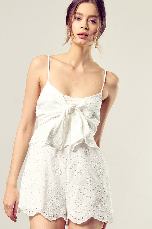 A woman wearing the Scallop Edge Front Tie-Up Romper in white, featuring a sleeveless design and eyelet texture, stands against a plain background, radiating boho style charm.