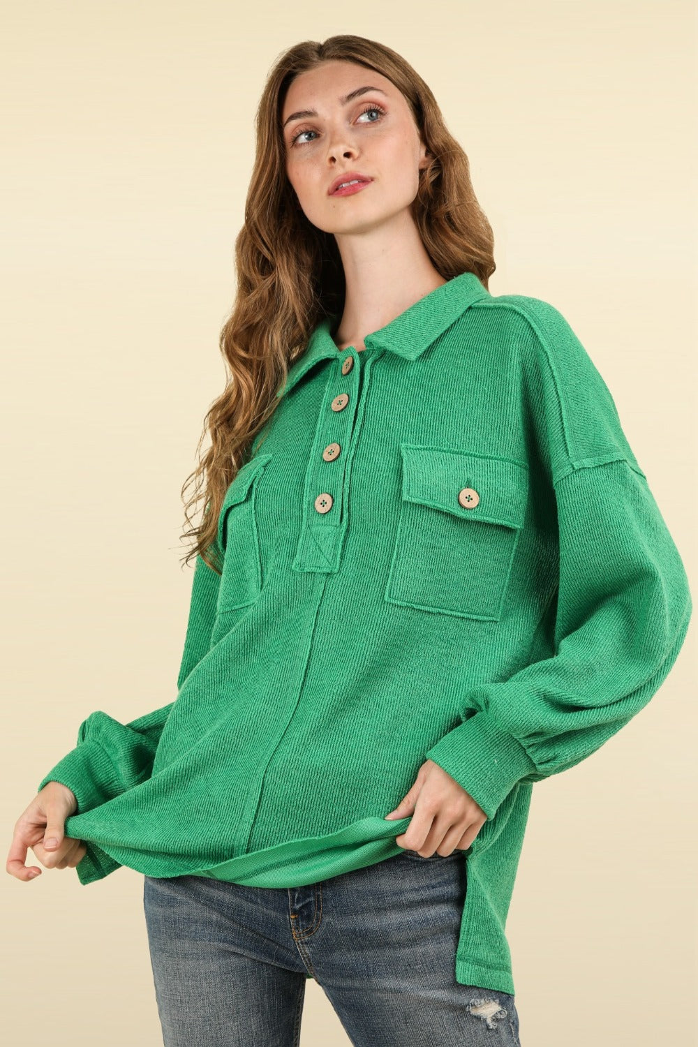 A person with long hair wearing an oversized fit, green VERY J Collared Half Button Knit Top with Pockets stands against a beige background.