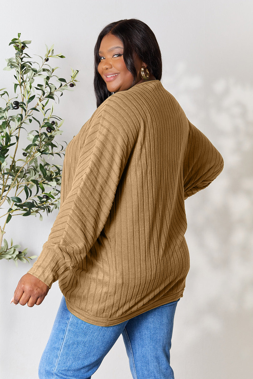 A person with wavy hair, wearing the Basic Bae Full Size Ribbed Cocoon Cardigan over a white top and beige pants, stands in front of a light-colored background with a leafy plant, showcasing an effortlessly basic style.