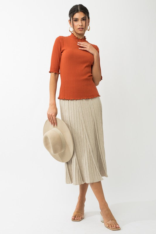 A person in a rust-colored Short Sleeve Mock Neck Merrow Edge Top holds a beige hat. They are also wearing a pleated skirt.