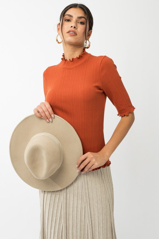 A person in a rust-colored Short Sleeve Mock Neck Merrow Edge Top holds a beige hat. They are also wearing a pleated skirt.