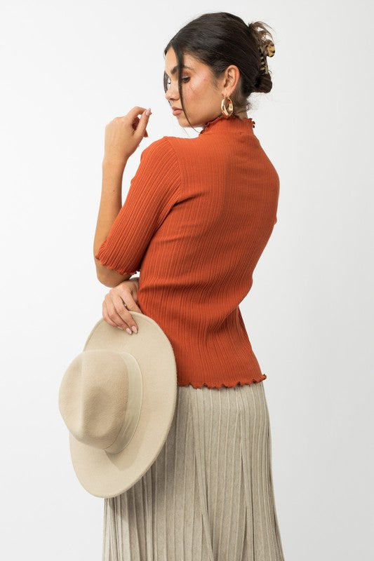 A person in a rust-colored Short Sleeve Mock Neck Merrow Edge Top holds a beige hat. They are also wearing a pleated skirt.