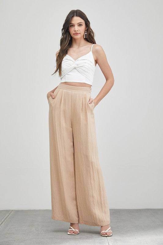 A woman stands against a plain backdrop, highlighting her summer fashion with the Front Twist Cami Top and a beige skirt. Her long brown hair falls elegantly, accentuated by stylish earrings. This versatile top adds a chic touch to her effortless ensemble.