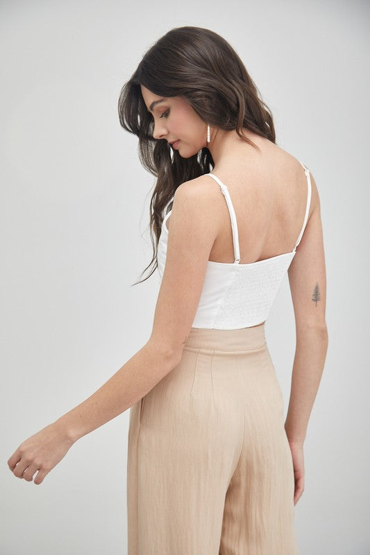 A woman stands against a plain backdrop, highlighting her summer fashion with the Front Twist Cami Top and a beige skirt. Her long brown hair falls elegantly, accentuated by stylish earrings. This versatile top adds a chic touch to her effortless ensemble.