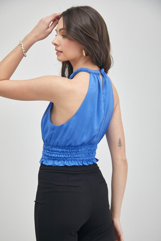 A woman wearing a chic, versatile wardrobe essential—a blue Smocked Waist Top paired with black pants—stands in front of a plain background, highlighting its elegant texture.