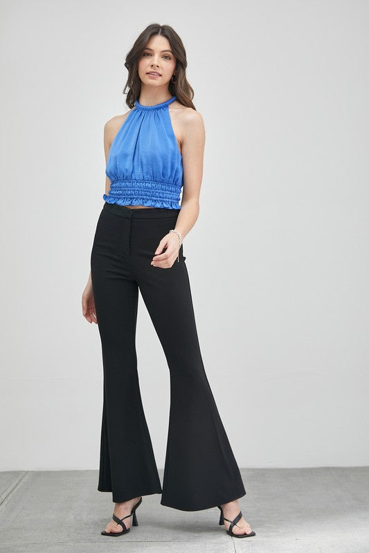 A woman wearing a chic, versatile wardrobe essential—a blue Smocked Waist Top paired with black pants—stands in front of a plain background, highlighting its elegant texture.