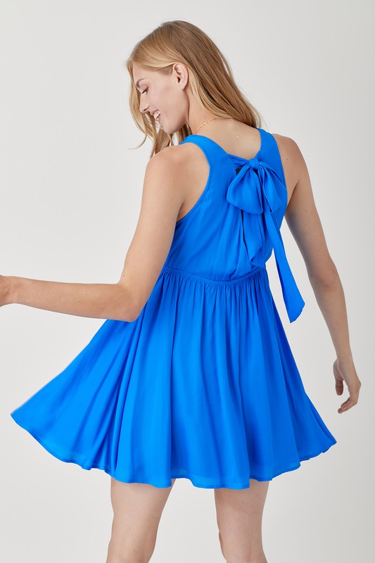 A woman wearing a relaxed fit V Neck Smock Sleeveless Dress in bright blue stands against a plain background, looking down with a slight smile.