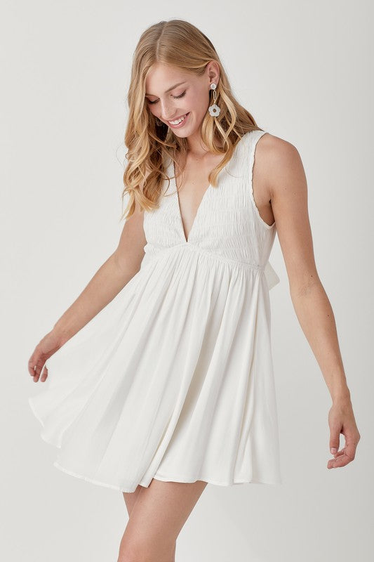 A person wearing the V Neck Smock Sleeveless Dress in white smiles while looking down, their hands gently holding the dress.