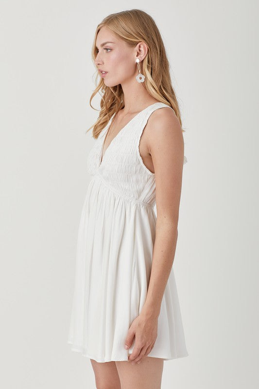 A person wearing the V Neck Smock Sleeveless Dress in white smiles while looking down, their hands gently holding the dress.