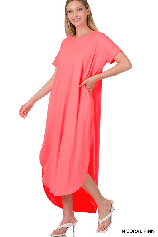 The person is glowing in a loose, coral pink Brushed DTY Short Sleeve Maxi Dress, paired elegantly with white sandals while standing and smiling with one hand casually tucked in the pocket.