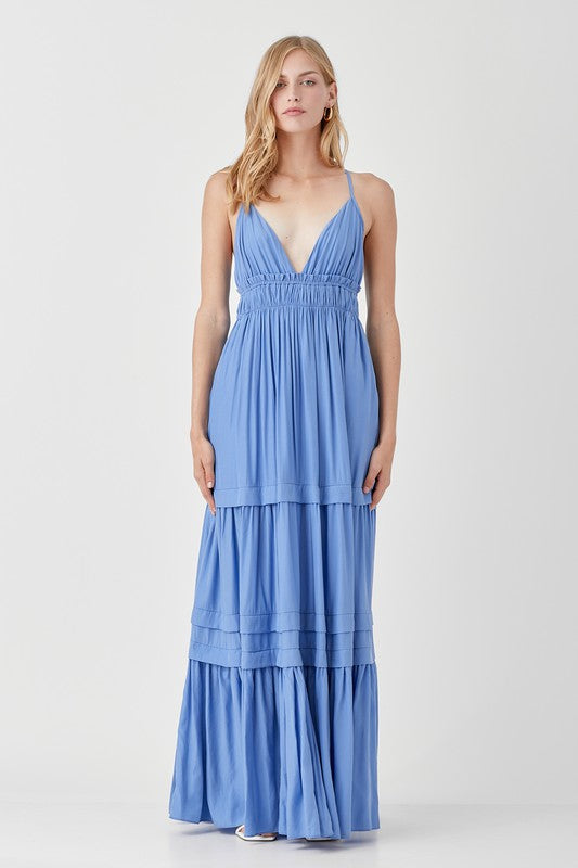 Dressed in a Shirred Ruffle Folded Detail Maxi Dress, featuring a striking blue color, plunging neckline, and stylish ruffle accents, the individual stands against a plain background exuding an effortless boho style.