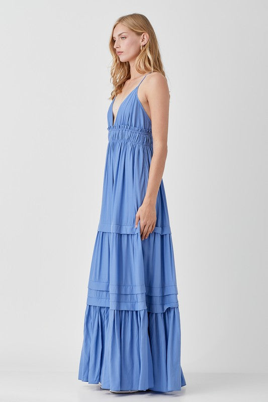 Dressed in a Shirred Ruffle Folded Detail Maxi Dress, featuring a striking blue color, plunging neckline, and stylish ruffle accents, the individual stands against a plain background exuding an effortless boho style.