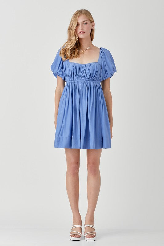 A person in a Pleated Front Criss Cross Back Strap Dress in blue, featuring short sleeves, stands smiling with one hand at their side against a neutral background.