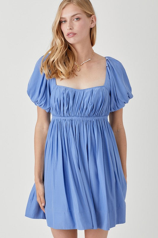 A person in a Pleated Front Criss Cross Back Strap Dress in blue, featuring short sleeves, stands smiling with one hand at their side against a neutral background.