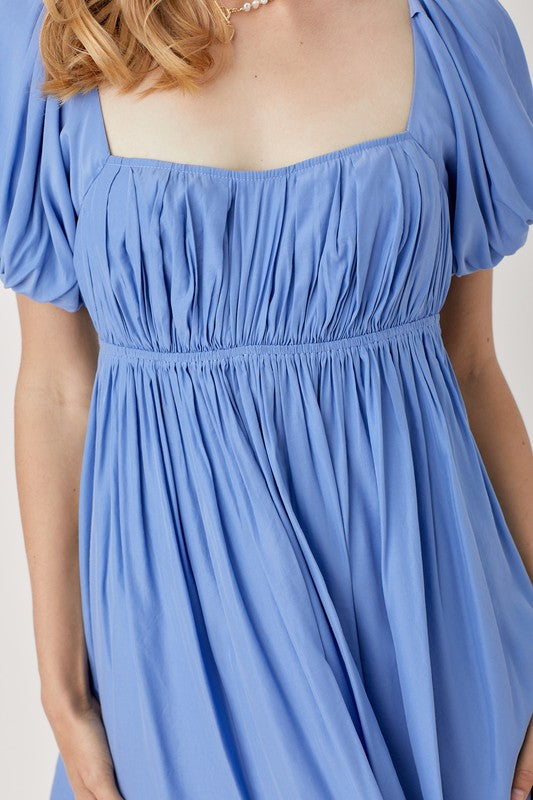 A person in a Pleated Front Criss Cross Back Strap Dress in blue, featuring short sleeves, stands smiling with one hand at their side against a neutral background.