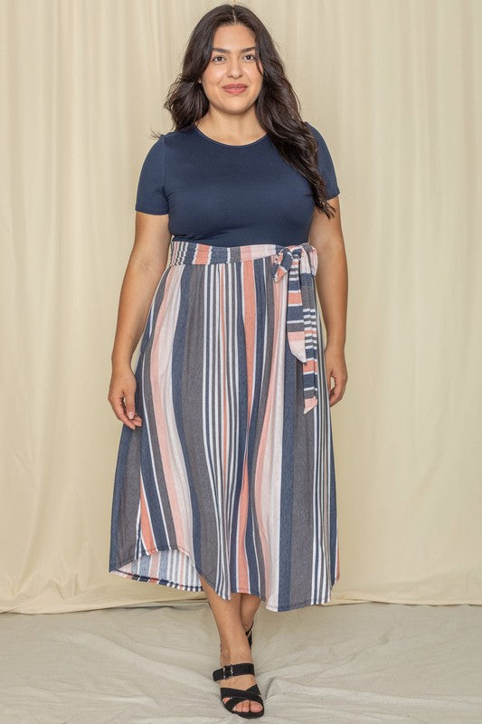 A woman in a Short Sleeve Vintage Sash Dress, featuring a navy top and striped skirt with a tie belt, stands gracefully in front of a beige curtain.