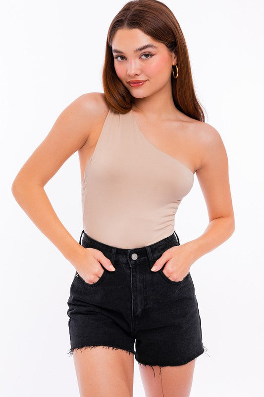 A person exudes chic style in a beige One Shoulder Bodysuit paired with fitted black denim shorts, posing elegantly against a white background.