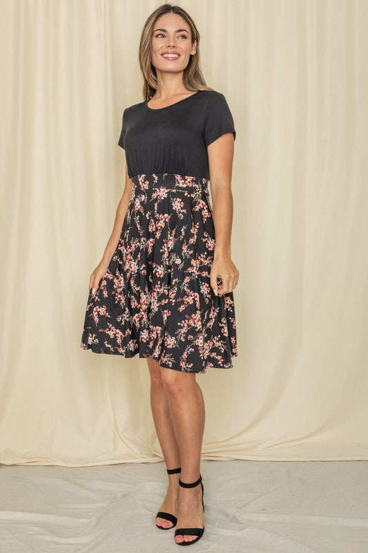 Wearing a Floral Band Flare Dress, a woman stands gracefully against a light-colored curtain, complemented by her black top.