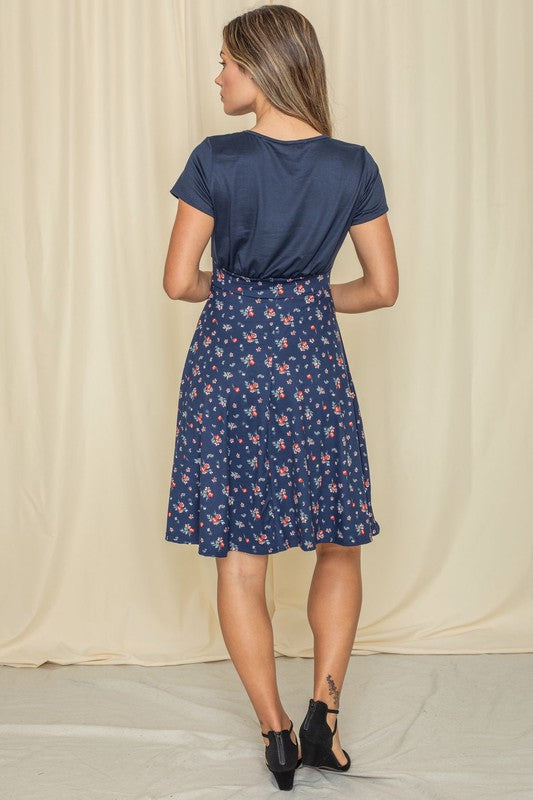 Wearing a Floral Band Flare Dress, a woman stands gracefully against a light-colored curtain, complemented by her black top.