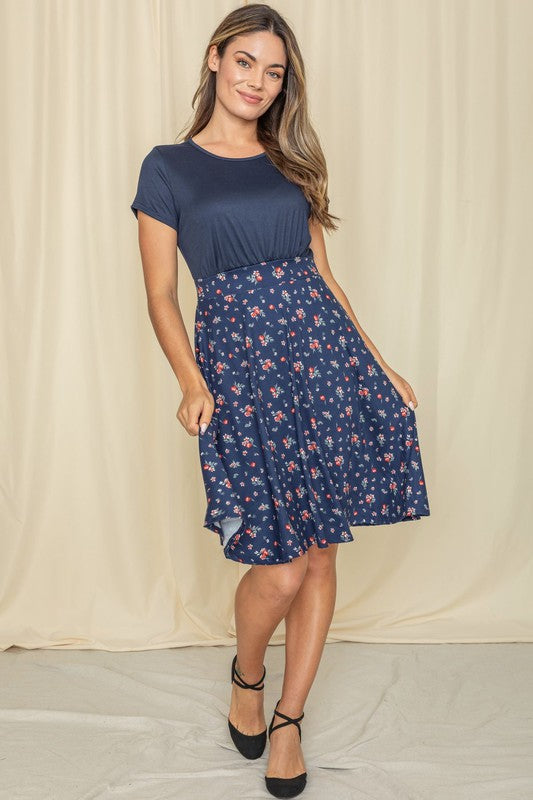 Wearing a Floral Band Flare Dress, a woman stands gracefully against a light-colored curtain, complemented by her black top.