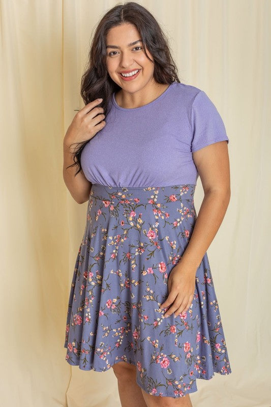 A woman in a vibrant purple top and a Plus Floral Band Flare Dress smiles against a light-colored background, proudly showcasing an outfit made in the United States.