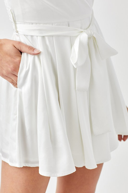 A woman stands against a plain background, looking forward while wearing the stylish Pleated Puff Sleeve Overwrap Romper in white.