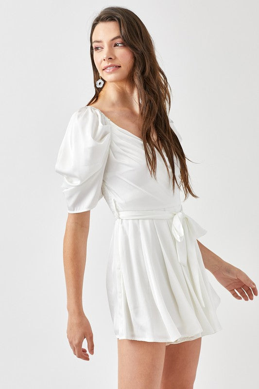 A woman stands against a plain background, looking forward while wearing the stylish Pleated Puff Sleeve Overwrap Romper in white.