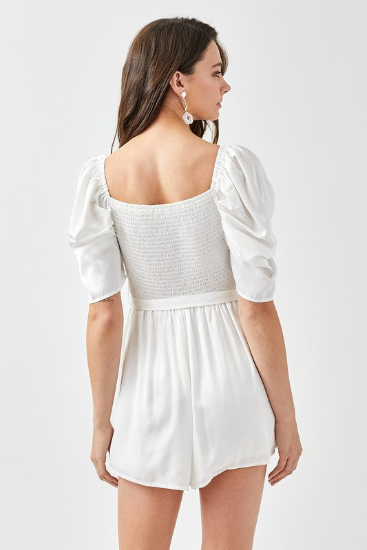 A woman stands against a plain background, looking forward while wearing the stylish Pleated Puff Sleeve Overwrap Romper in white.