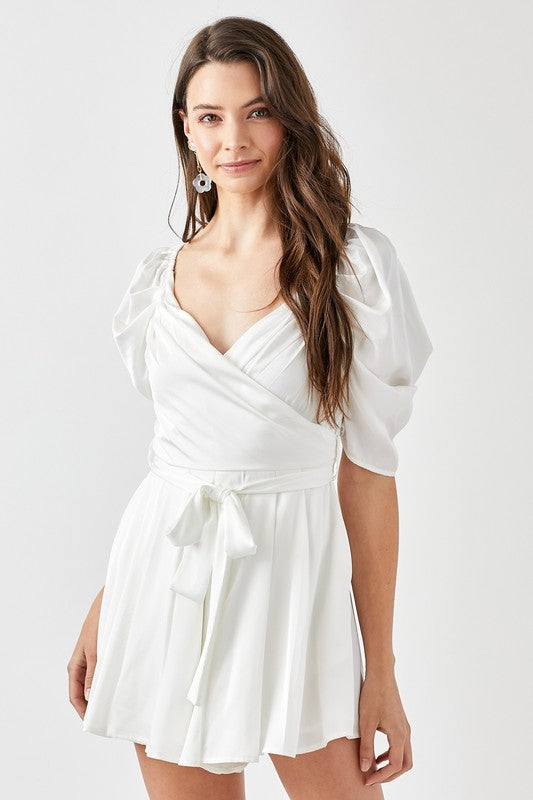 A woman stands against a plain background, looking forward while wearing the stylish Pleated Puff Sleeve Overwrap Romper in white.