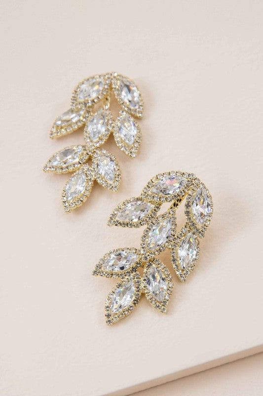 Lana Drop Earrings, showcasing leaf-shaped crystal designs set in a gold petaled frame, displayed on a beige surface.