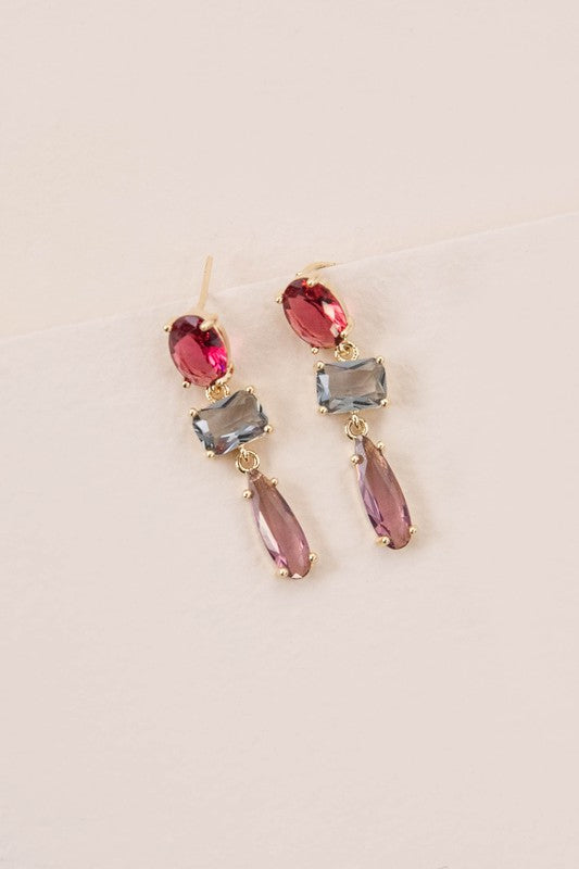 The Fraise Dangle Earrings feature red and blue rectangular, as well as purple teardrop gemstones, elegantly arranged on a white surface. With 10k gold plating, these stunning earrings are perfect for adding a touch of formal style to any outfit.