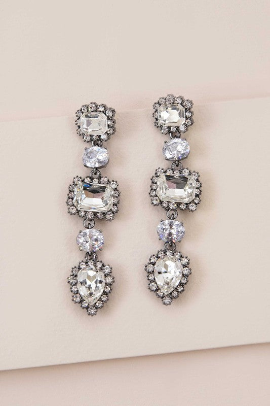 The Regal Drop Earrings are an ornate piece crafted from sterling silver, featuring an array of rectangular, circular, and teardrop-shaped rhinestone jewels.