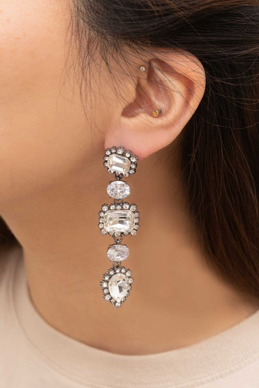 The Regal Drop Earrings are an ornate piece crafted from sterling silver, featuring an array of rectangular, circular, and teardrop-shaped rhinestone jewels.