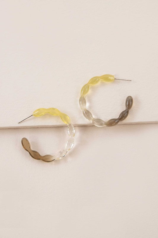 The Jelly Hoop Earrings, featuring translucent acrylic hoops with wavy designs in yellow, gray, and a subtle third tone, rest elegantly on a textured surface. Equipped with sterling silver posts, they provide both style and comfort.