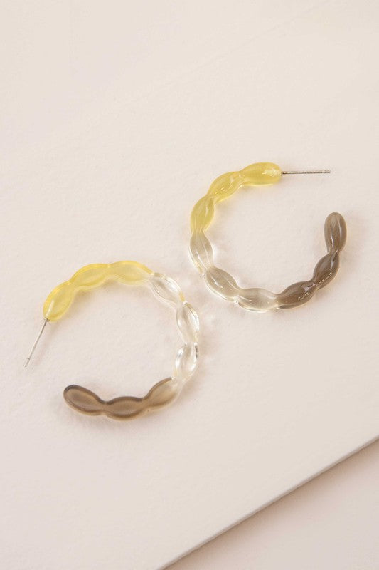 The Jelly Hoop Earrings, featuring translucent acrylic hoops with wavy designs in yellow, gray, and a subtle third tone, rest elegantly on a textured surface. Equipped with sterling silver posts, they provide both style and comfort.