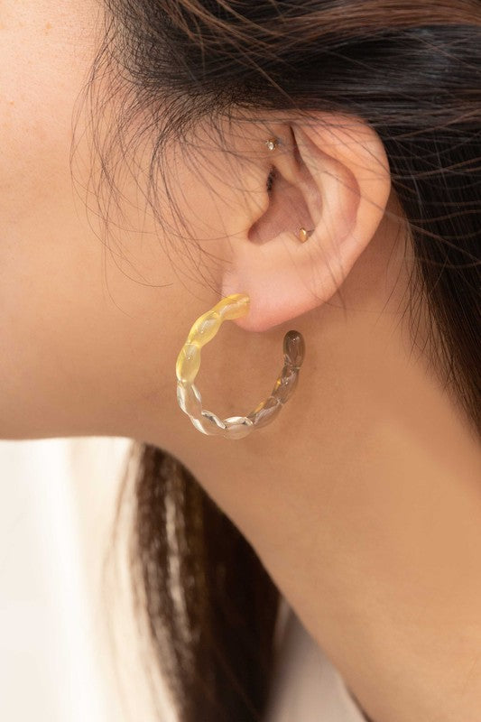 The Jelly Hoop Earrings, featuring translucent acrylic hoops with wavy designs in yellow, gray, and a subtle third tone, rest elegantly on a textured surface. Equipped with sterling silver posts, they provide both style and comfort.