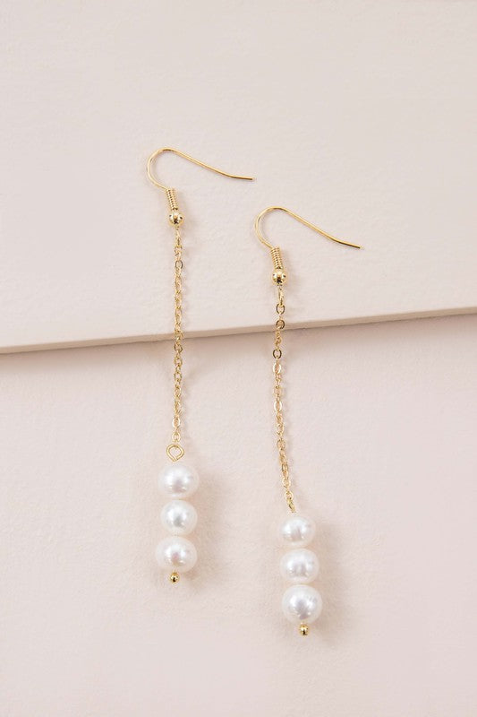 The Ines Pearl Hook Earrings showcase three lustrous freshwater pearls elegantly suspended from a delicate chain, designed in gold against a light background.