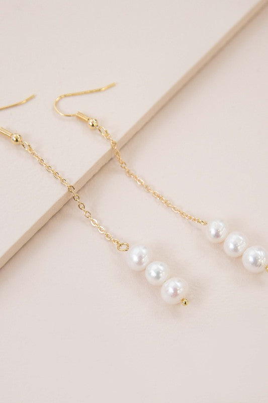 The Ines Pearl Hook Earrings showcase three lustrous freshwater pearls elegantly suspended from a delicate chain, designed in gold against a light background.