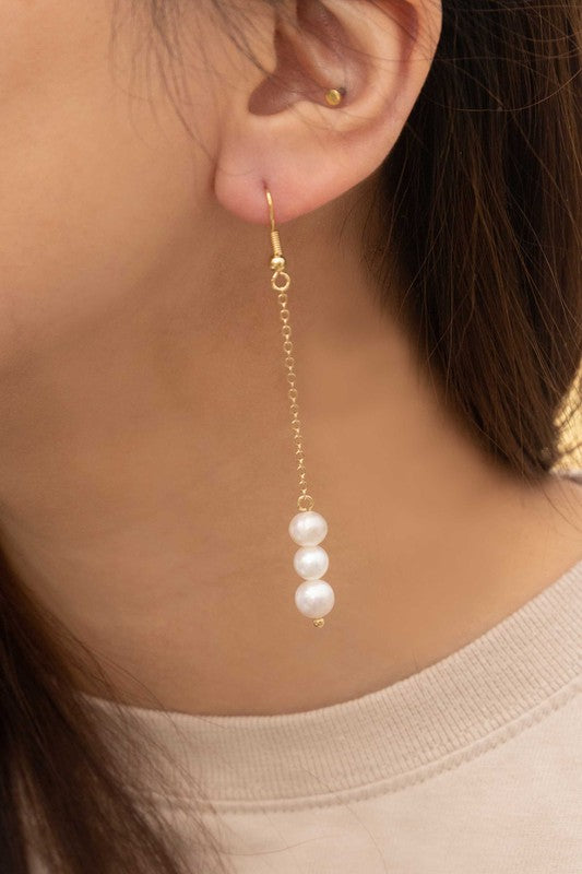 The Ines Pearl Hook Earrings showcase three lustrous freshwater pearls elegantly suspended from a delicate chain, designed in gold against a light background.