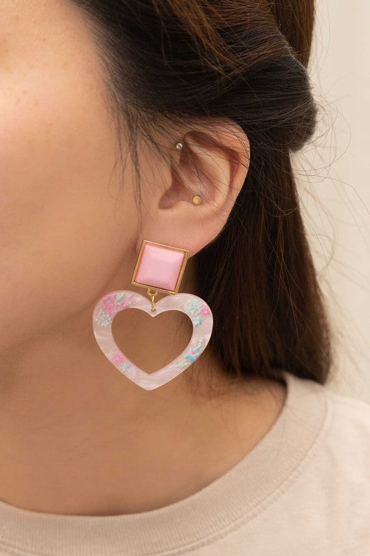 The Lucy Heart Earrings, radiating the charm of a 2000s pop star, highlight pink and floral accents combined with square pink studs set against a white background. These earrings are complemented by a sterling silver post for an extra touch of elegance.