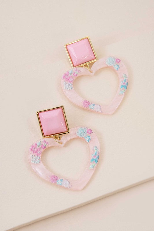 The Lucy Heart Earrings, radiating the charm of a 2000s pop star, highlight pink and floral accents combined with square pink studs set against a white background. These earrings are complemented by a sterling silver post for an extra touch of elegance.