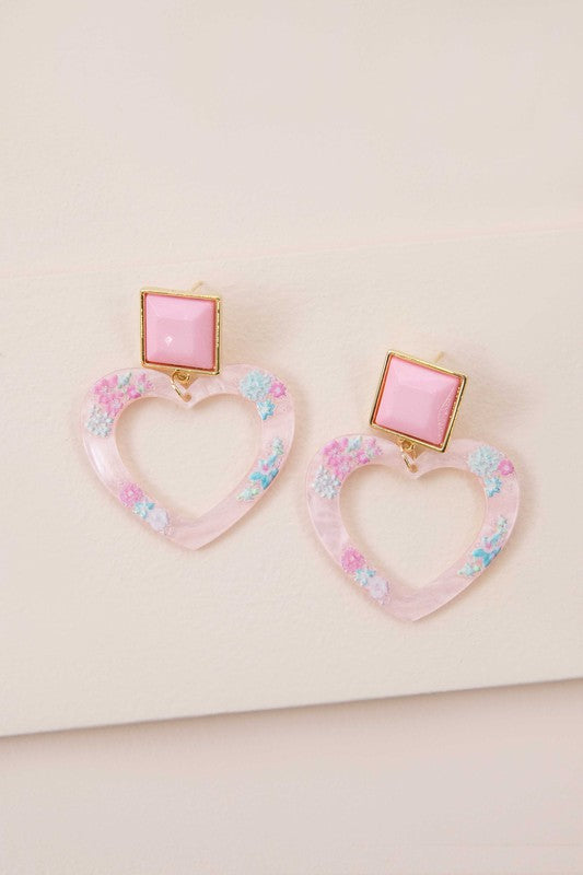 The Lucy Heart Earrings, radiating the charm of a 2000s pop star, highlight pink and floral accents combined with square pink studs set against a white background. These earrings are complemented by a sterling silver post for an extra touch of elegance.