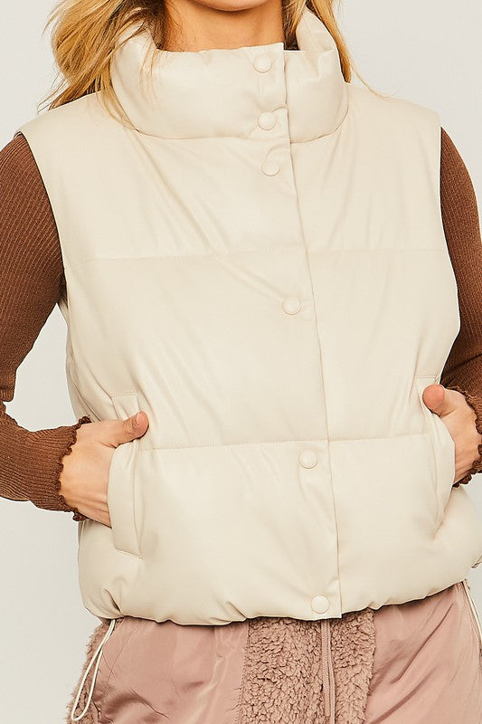 A woman stands against a plain background, dressed in the PU Padded Vest, which is beige with button embellishments. She pairs it with a brown long-sleeve top and pink fleece pants.