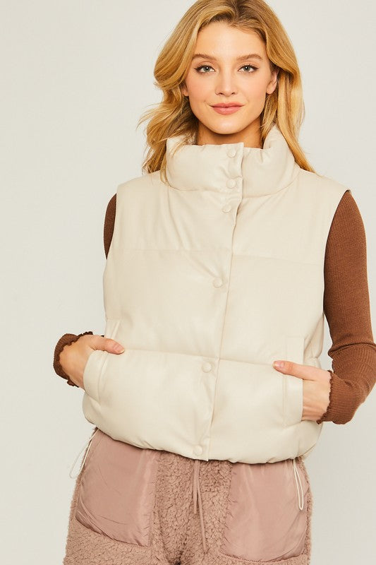 A woman stands against a plain background, dressed in the PU Padded Vest, which is beige with button embellishments. She pairs it with a brown long-sleeve top and pink fleece pants.