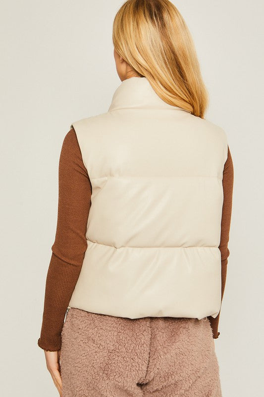 A woman stands against a plain background, dressed in the PU Padded Vest, which is beige with button embellishments. She pairs it with a brown long-sleeve top and pink fleece pants.