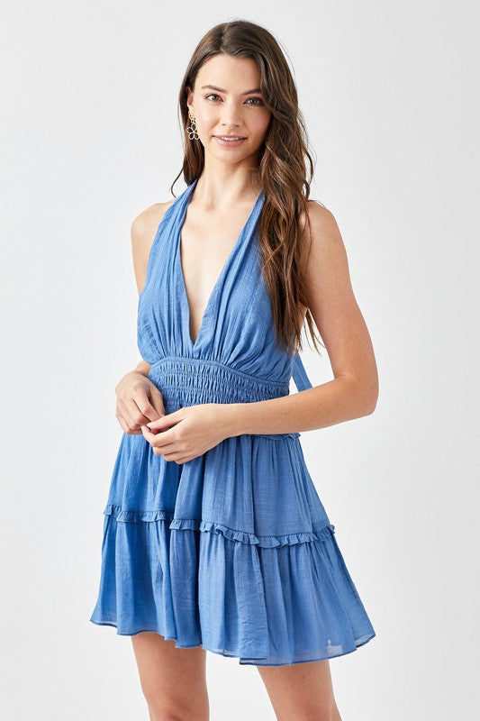 A woman showcases the stylish Halter Neck Smocked Ruffle Dress in blue, gracefully posing with one hand on her hip against a plain backdrop.