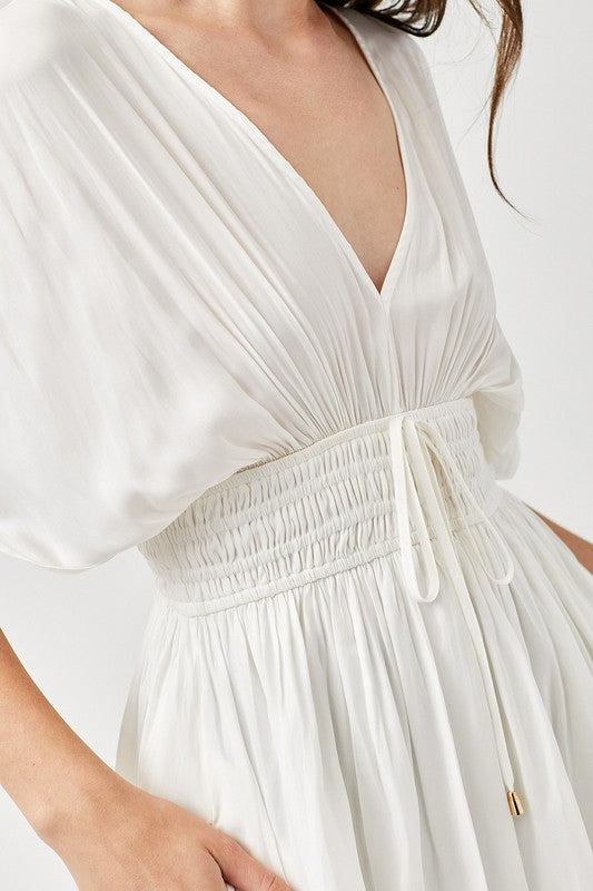 A woman is wearing a Smocked Waist with Tassel Strap Dress, featuring a white color, short sleeves, and a V-neckline, as she stands against a plain background.