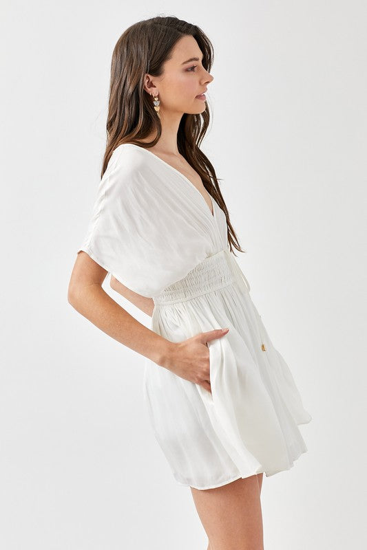 A woman is wearing a Smocked Waist with Tassel Strap Dress, featuring a white color, short sleeves, and a V-neckline, as she stands against a plain background.