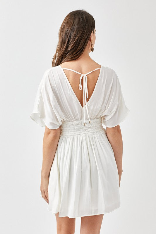 A woman is wearing a Smocked Waist with Tassel Strap Dress, featuring a white color, short sleeves, and a V-neckline, as she stands against a plain background.